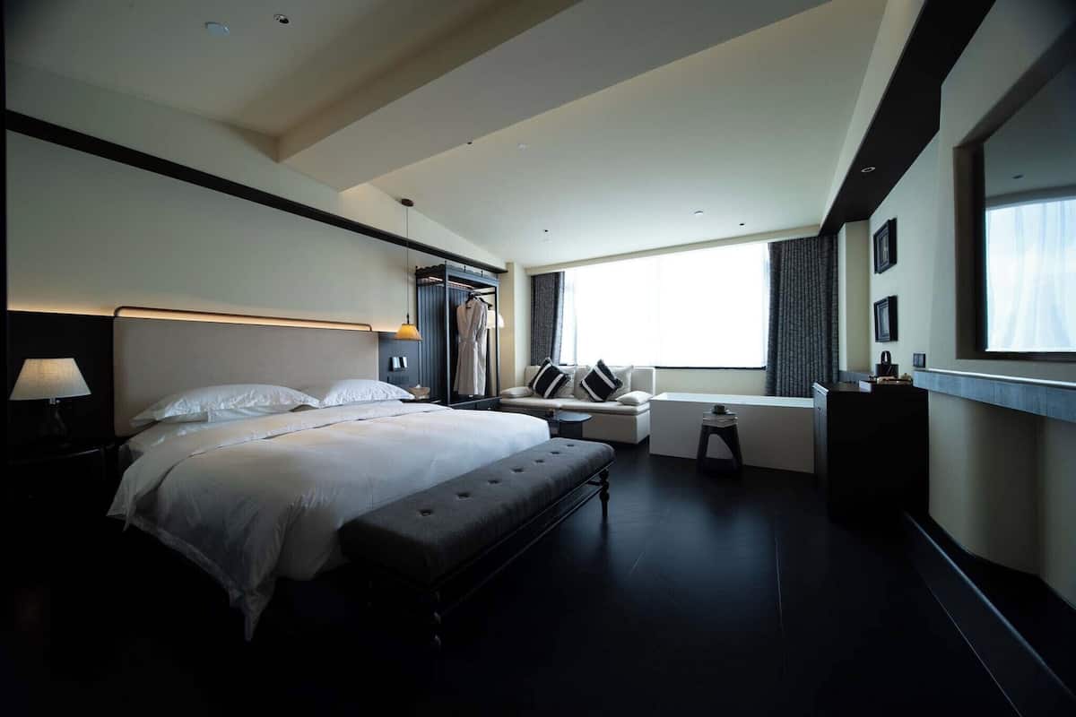 A modern hotel room with a large bed, bedside tables, a sofa, a small table, and a window with curtains.