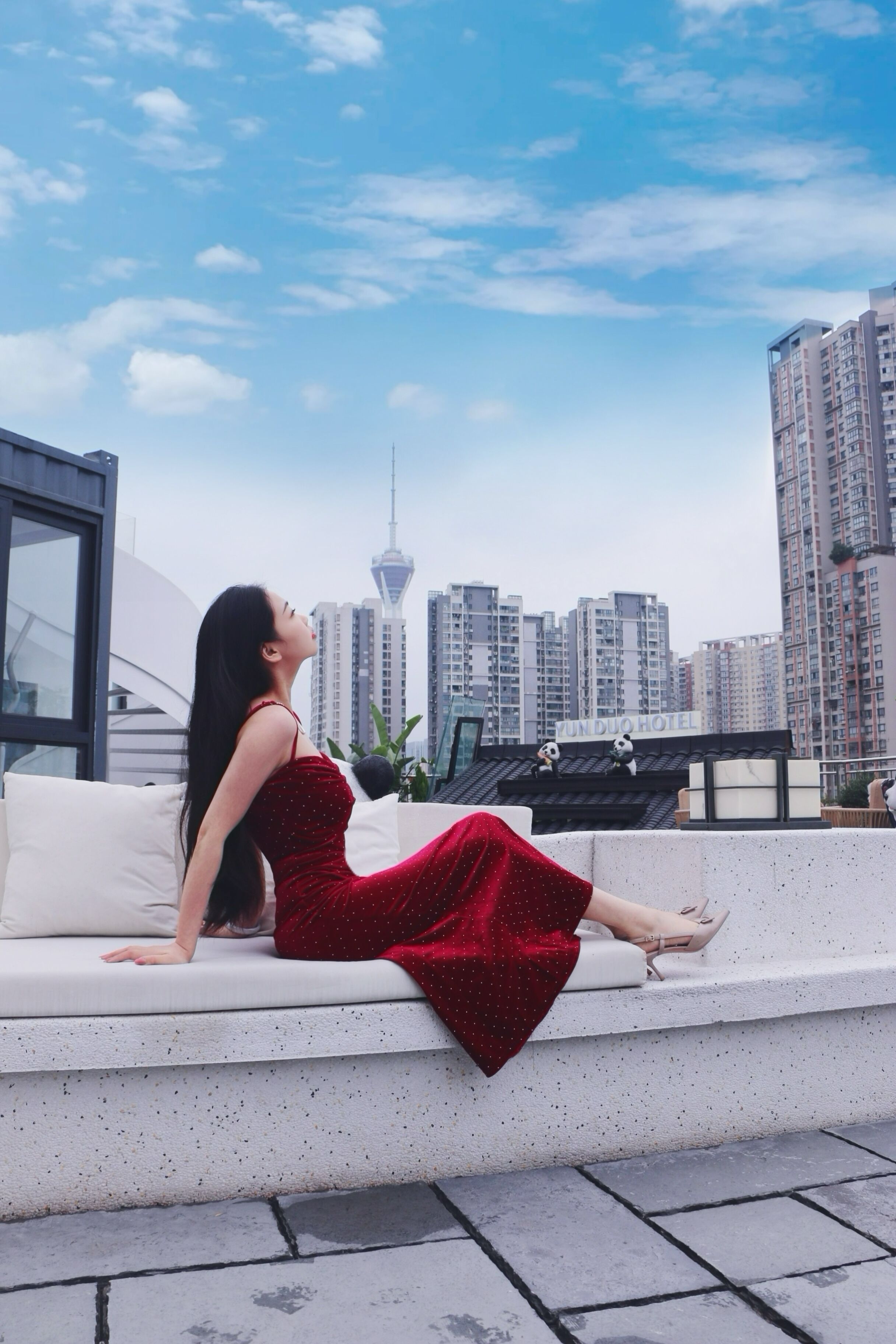 Una mujer con un vestido rojo sentada en un sofá blanco al aire libre con vistas a un paisaje urbano, incluyendo una torre y edificios altos.