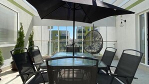 A glass-top table with four black chairs, a large black umbrella, and a hanging spherical light fixture on a balcony with a clear view of buildings.