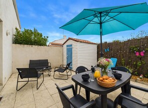 Eine Terrasse mit Tisch, Stühlen, einem blauen Sonnenschirm und einem kleinen Schuppen im Hintergrund.
