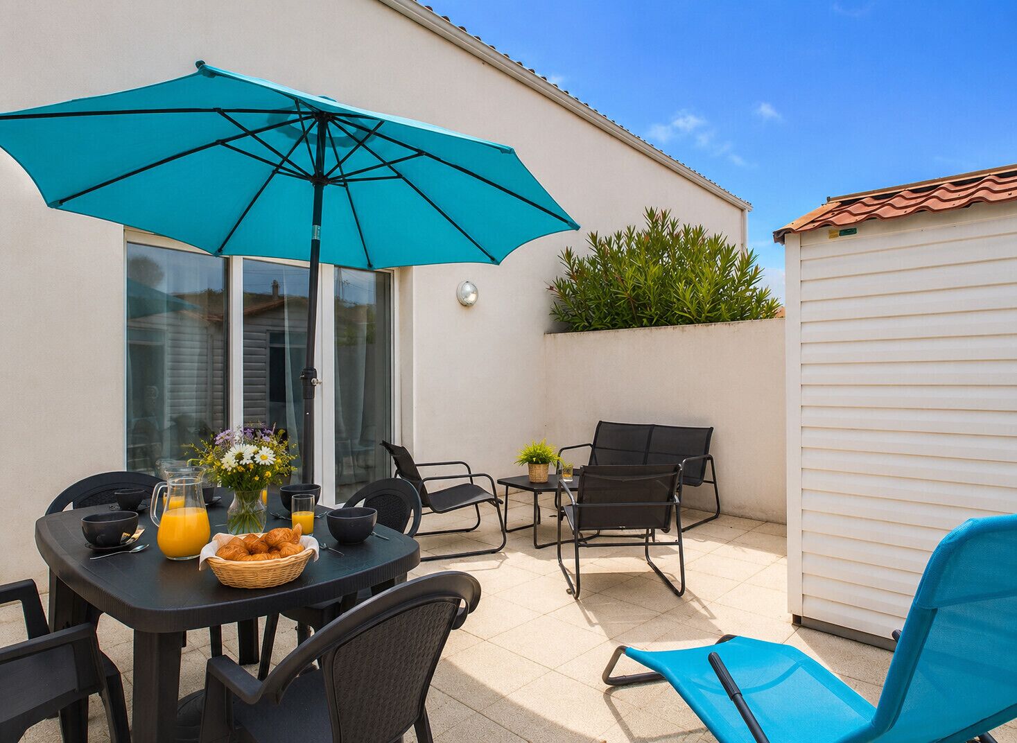 Um pátio com mesa posta para o café da manhã, cadeiras de exterior, um guarda-sol azul e uma pequena área de jardim.