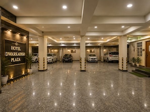 A hotel entrance with a sign reading 'HOTEL DWARKADISH PLAZA,' a parking area with several cars, and a clean, well-lit interior. - Hotel Dwarkadhish Plaza (Ayodhya)