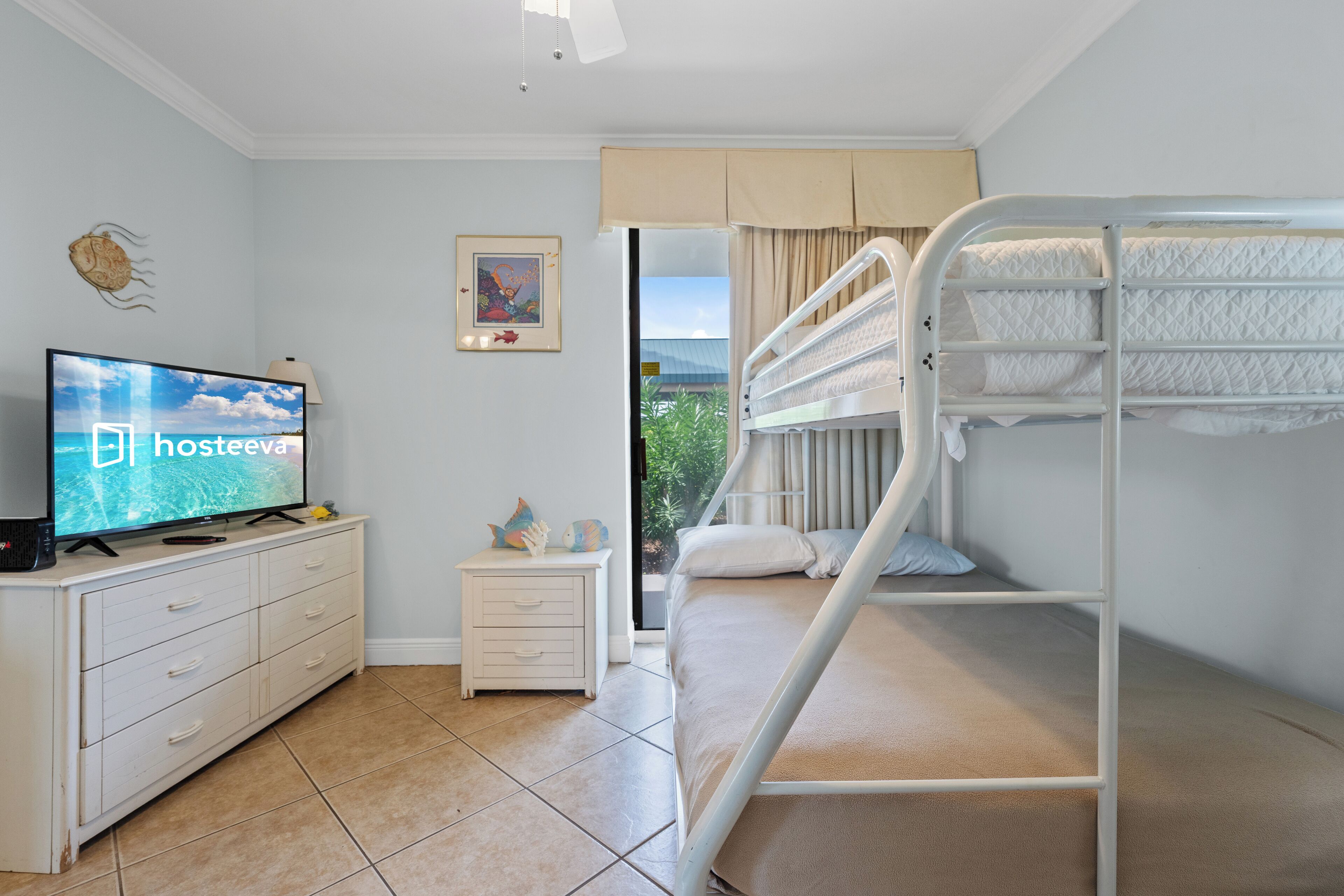 Une chambre avec un lit superposé, un téléviseur posé sur une commode, une petite table de chevet et une fenêtre donnant sur un paysage verdoyant.