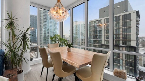 A modern dining area with a wooden table and chairs, large windows, and a hanging light fixture. - King Bed | Mins to Stampede | Cgy Twr View | UG Pk (Calgary)
