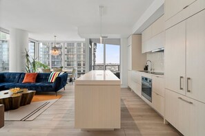 A modern apartment with a central island kitchen, a blue sofa, and large windows offering city views. - King Bed | Mins to Stampede | Cgy Twr View | UG Pk (Calgary)
