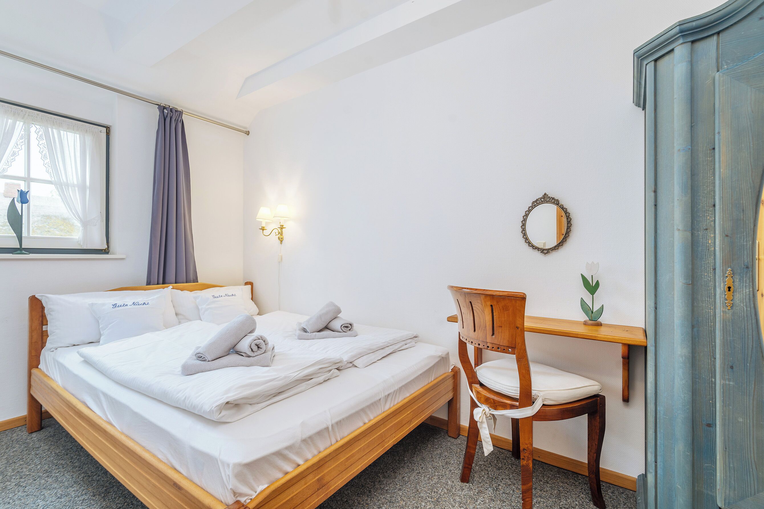 Un lit bien fait avec des oreillers, une chaise en bois, un bureau, un miroir fixé au mur et une fenêtre avec des rideaux.