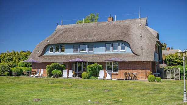 Ein Gebäude mit einem Reetdach und einem Backsteinunterbau, Außenbestuhlung mit Sonnenschirmen und einem gepflegten Rasen.