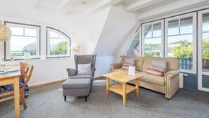 Ein gemütliches Zimmer mit hohen Decken, einer Couch, einem Sessel, einem kleinen Tisch und großen Fenstern mit Blick ins Freie.