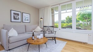 A living room with a sofa, armchair, round coffee table, and a window with curtains. - Modern living under thatch - your stylish Bodden break for two (Born a. Darß)