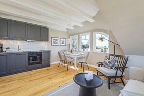 A modern kitchen with grey cabinets, a dining area with a white table and chairs, and a cozy seating area with a black chair and a round table. - Contemporary comfort under thatch - your maisonette on the Bodden (Born a. Darß)