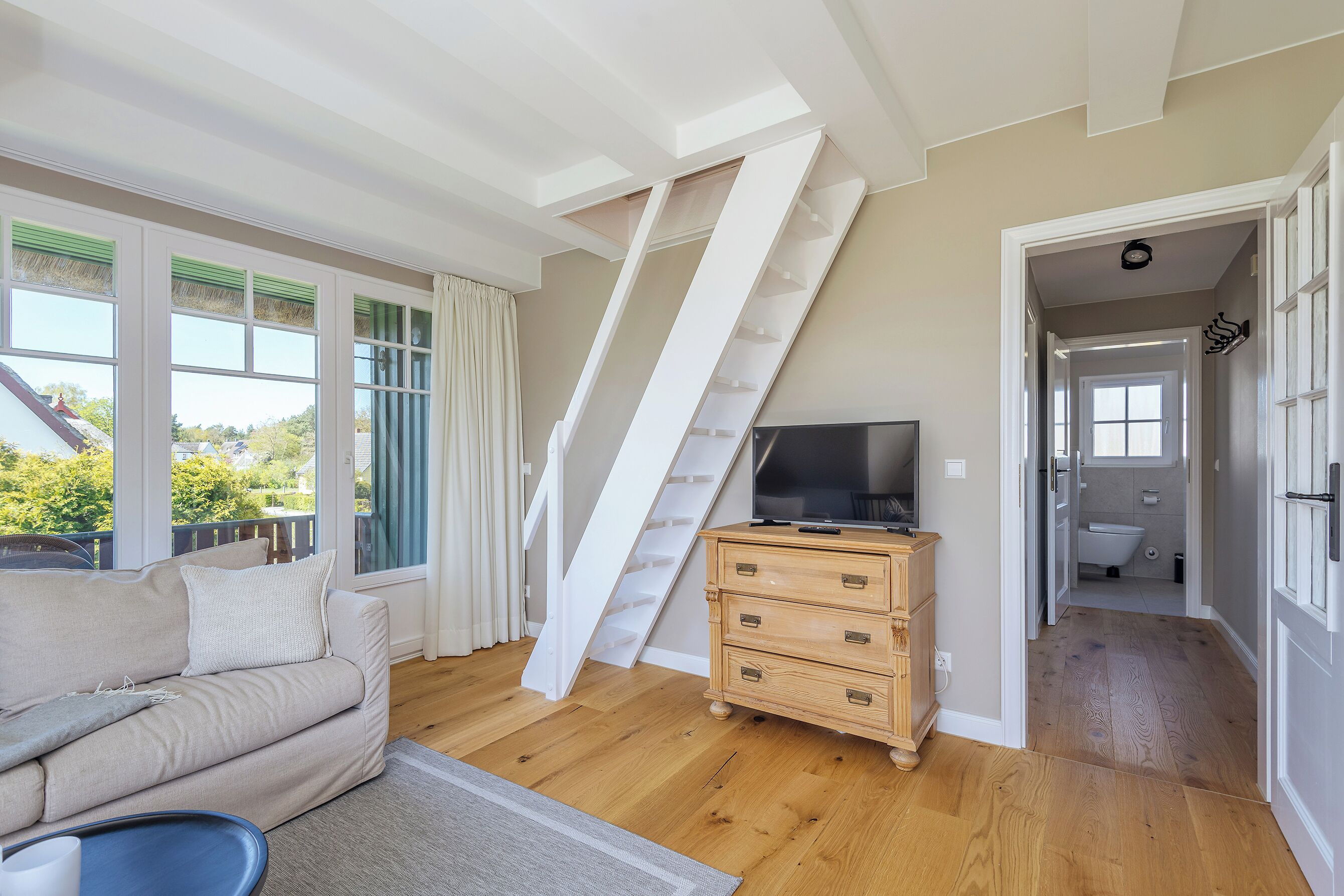 A room with a sofa, a TV on a wooden dresser, a staircase, and a bathroom visible through an open door.