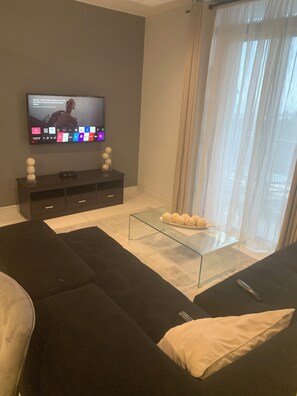 Una sala de estar moderna con un televisor de pantalla plana montado en la pared, una mesa de centro de vidrio con objetos decorativos, un sofá oscuro y un ventanal con cortinas translúcidas.