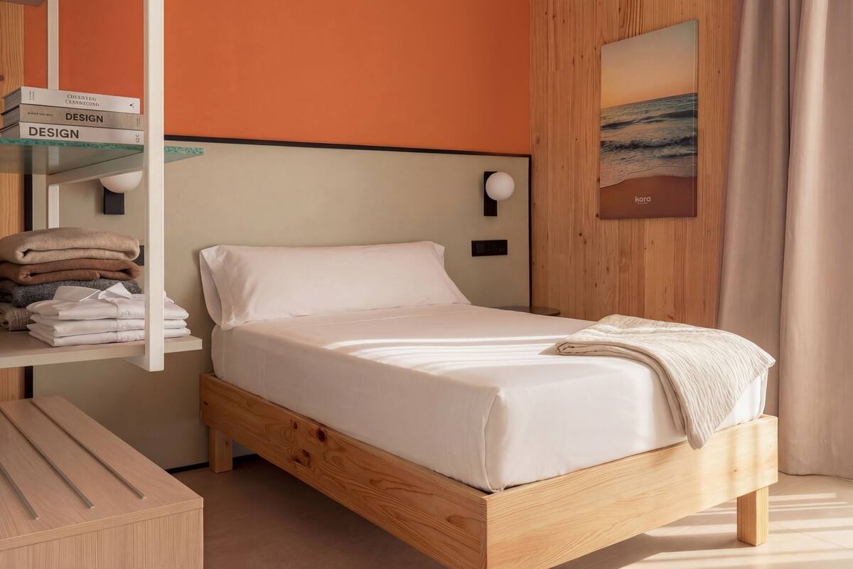 A neatly made bed in a modern room with a wooden headboard, a shelf with folded towels, and a painting of a beach scene on the wall.