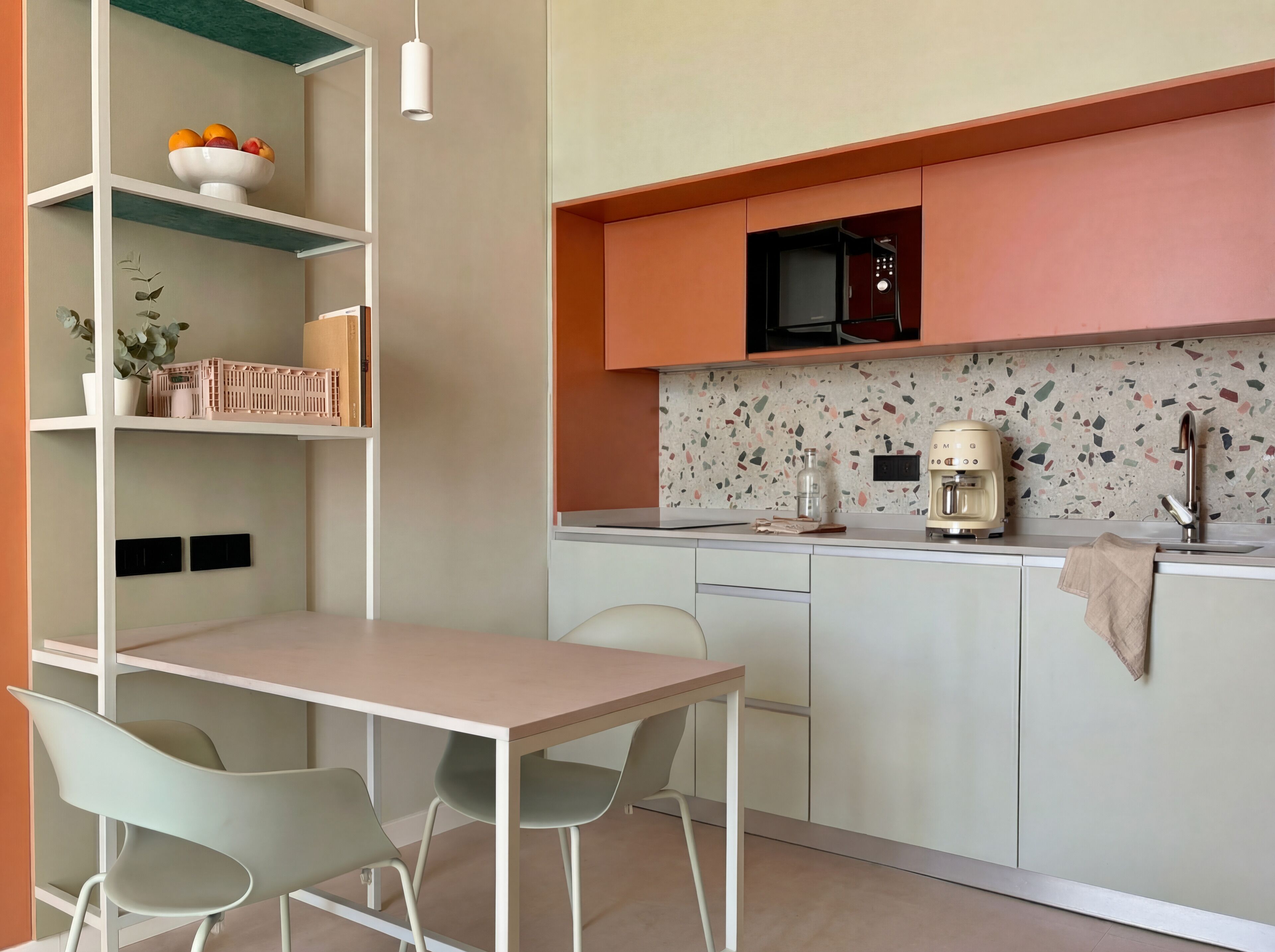 Een moderne keuken met witte kasten, een magnetron, koffiezetapparaat en een spoelbak. Er is een eettafel met stoelen en een plank met een fruitschaal.
