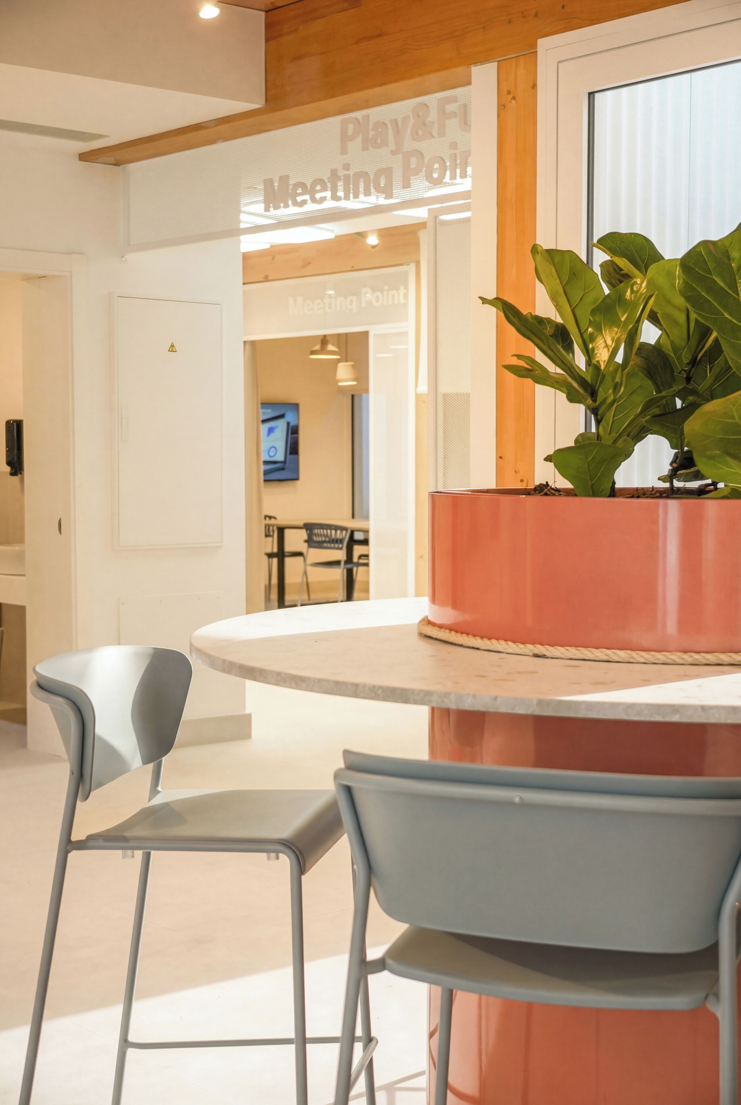 Un intérieur moderne avec une table ronde, deux chaises grises, une plante en pot et un panneau indiquant « Lieu de rencontre pour jouer et s’amuser ».