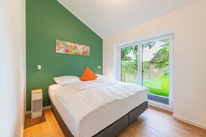 Ein Schlafzimmer mit einem Bett, einem Nachttisch, einer grünen Wand, einem Gemälde und Blick auf einen Garten durch eine Schiebetür.