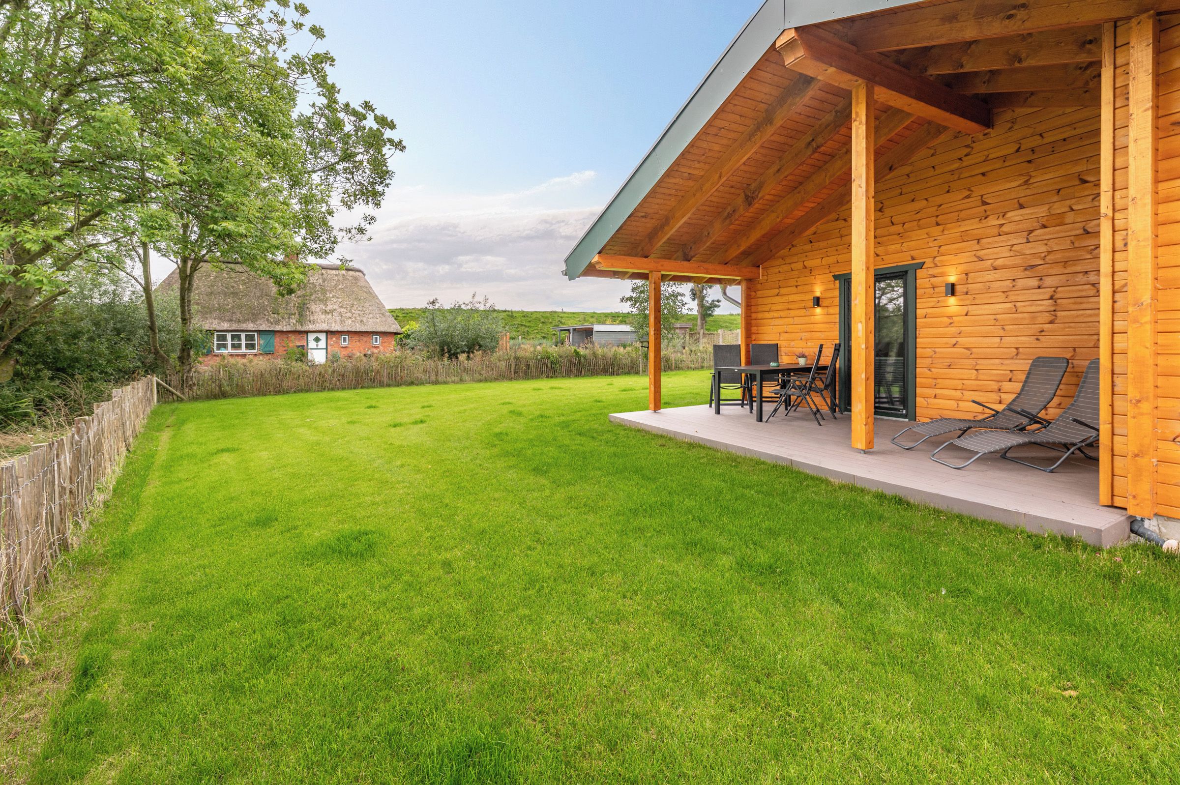 A wooden house with a covered patio, a lawn, and a traditional thatched-roof house in the background.