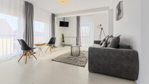 A modern living room with a grey sofa, a coffee table, and two chairs. There is a TV mounted on the wall, a floor lamp, and a large window with curtains. - MoHost Harmony Apartments - City Center  (Brașov)