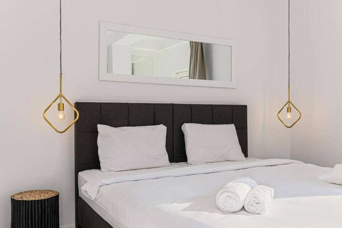 Una cama bien hecha con sábanas y almohadas blancas, un cabecero oscuro y dos lámparas colgantes con marco dorado.