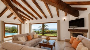 A spacious living room with a large window overlooking a pool and ocean, wooden beams on the ceiling, a fireplace, and a flat-screen TV mounted on the wall. - Charming rustic villa with sea views in Playa del Faro Camarinal (Tarifa)