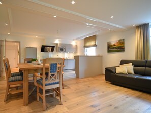 Ein modernes Wohnzimmer mit Esstisch aus Holz, Stühlen, einer Couch und einer Küche im Hintergrund.