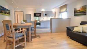 Cocina moderna con armarios blancos, microondas y televisor.  Comedor con mesa y sillas de madera, y sofá de cuero negro en la sala.