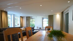 Una sala de estar moderna con una mesa de comedor de madera, sillas, un sofá y ventanas grandes con cortinas.