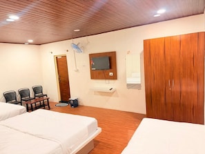 A hotel room with two beds, a wooden wardrobe, a TV mounted on the wall, a small table with chairs, and a ceiling fan. - AETHERIA ESTATE RETREAT (Chikkamagaluru)