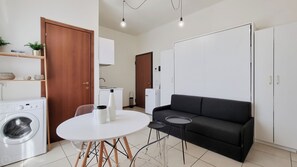 Ein kompakter Wohnraum mit Waschmaschine, einer schwarzen Couch, einem runden Esstisch und einem weißen Schrank.