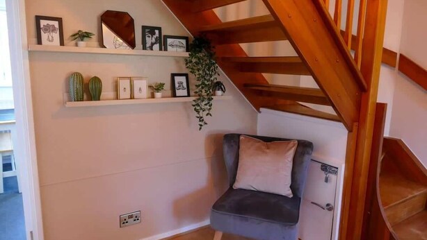 A room with a grey chair, wooden staircase, white walls, and shelves with framed pictures and plants. - Home In Longwell Green  Inbetween Bristol and Bath (Longwell Green)