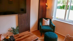 A living room with a flat-screen TV mounted on the wall, a green armchair with a cushion, a wooden coffee table, and a window with a view of greenery outside. - Home In Longwell Green  Inbetween Bristol and Bath (Longwell Green)