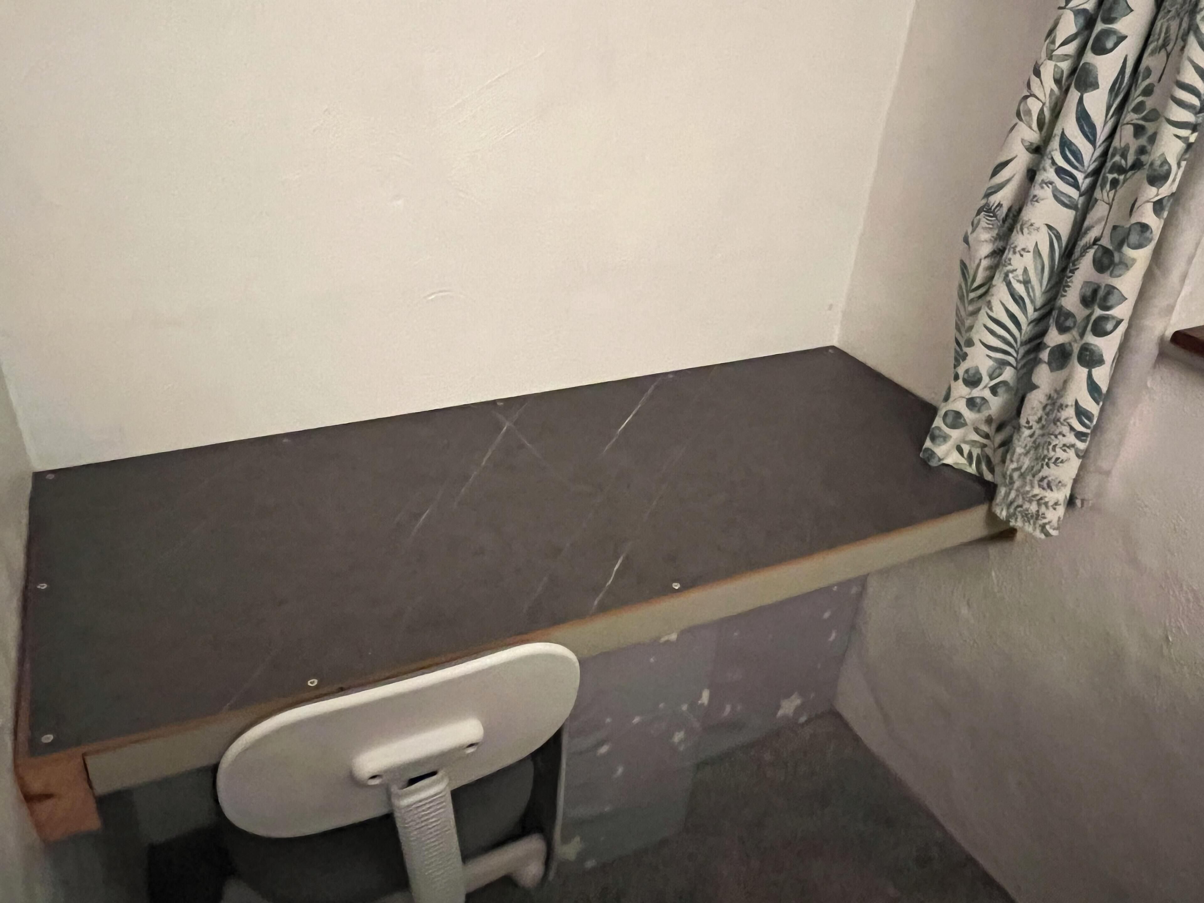 A small, dark-colored desk with a white chair in a corner of a room with plain walls and a curtain with a leaf pattern.