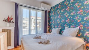 Ett hotellrum med en stor säng, vita sängkläder, en blommig tapet och utsikt över byggnader genom fönstret.