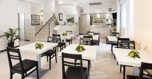 Un comedor moderno con mesas blancas y sillas negras, que incluye una escalera y una cocina al fondo.