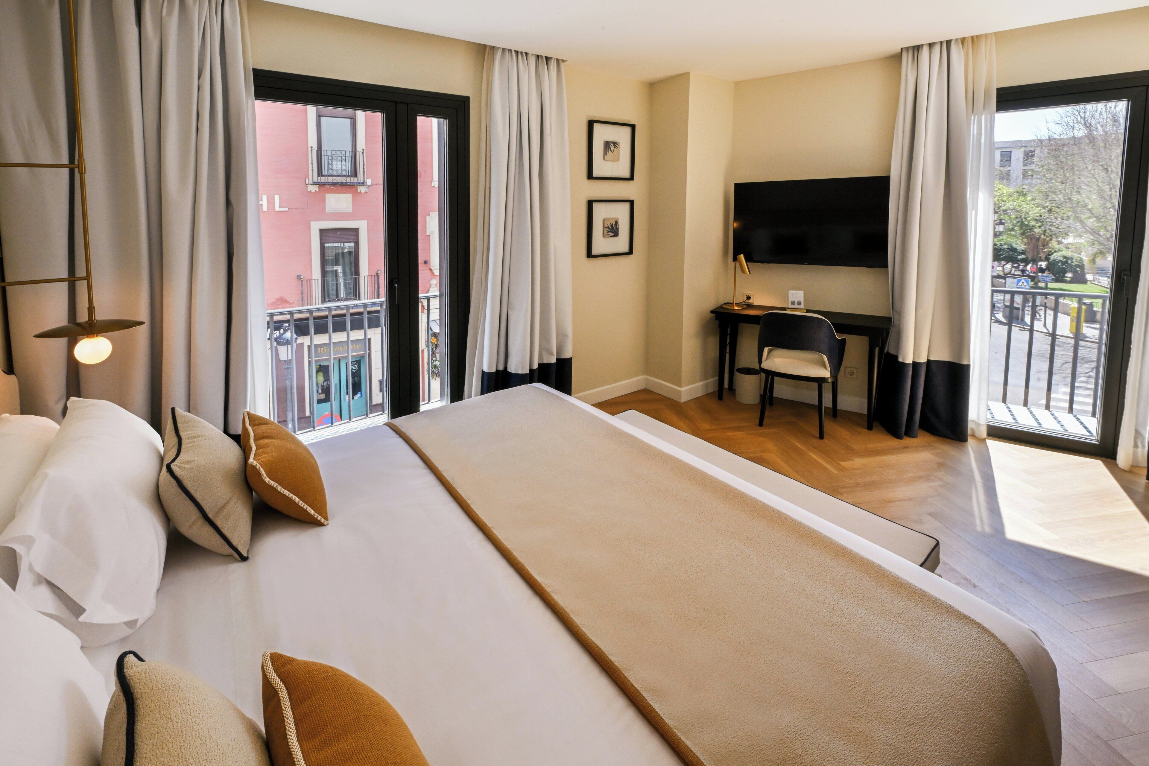 A hotel room with a large bed, a desk, a chair, a TV, and a balcony with a view of buildings.