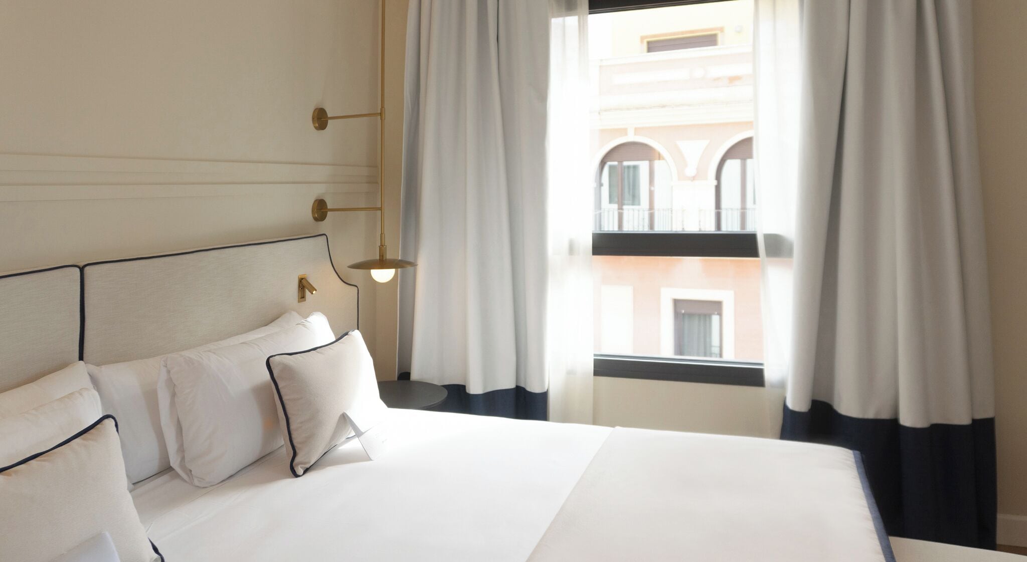 Ein Hotelzimmer mit einem großen Bett, einem Kopfteil mit einer Leuchte und Blick auf ein Gebäude durch das Fenster.