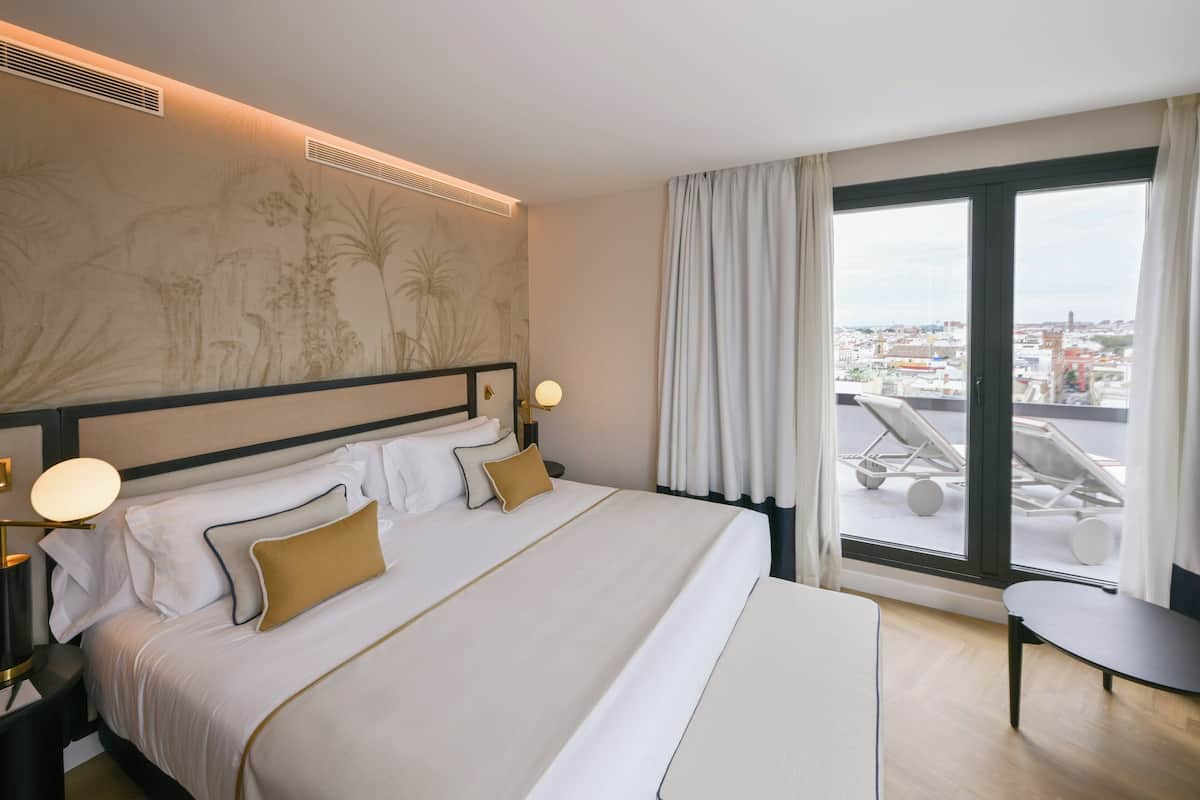 A hotel room with a large bed, a view of the city through a large window, and a small table.