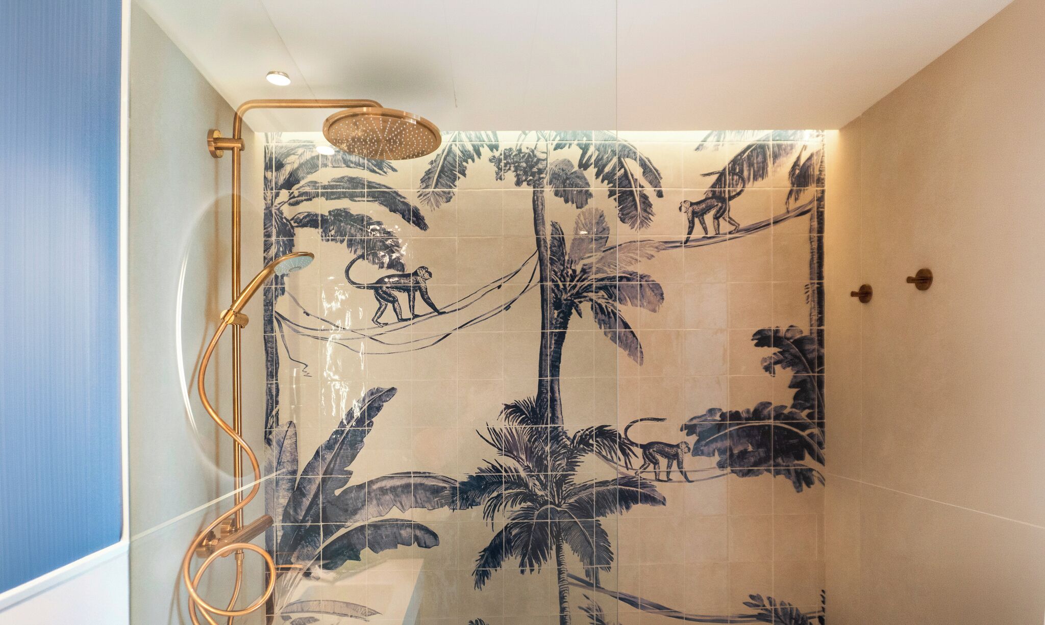 Ducha con cabezal dorado y tipo lluvia, rodeada de azulejos con motivos de palmeras.