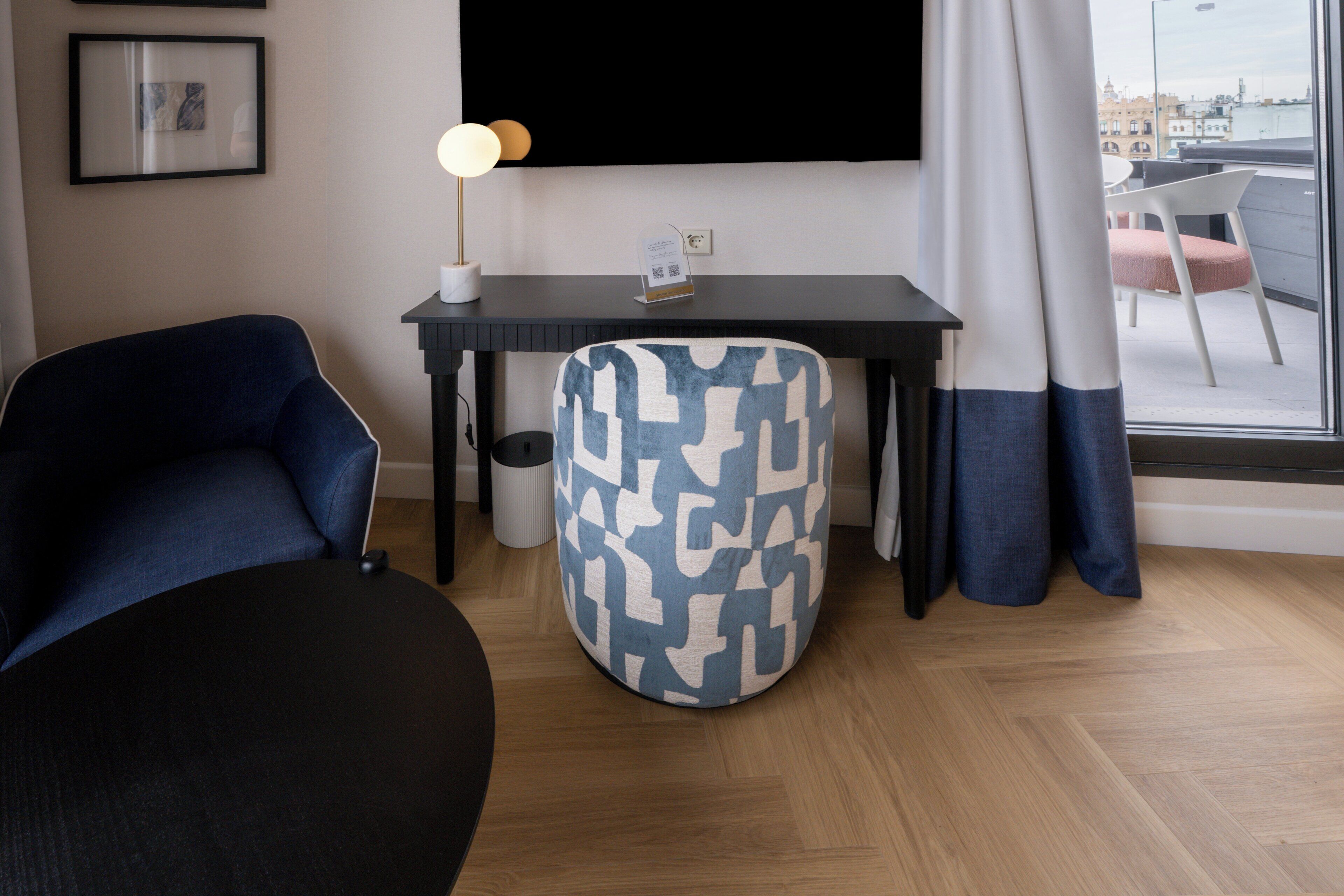 A modern room with a black desk, a round blue and white patterned ottoman, a dark blue sofa, and a view of the city through a large window.