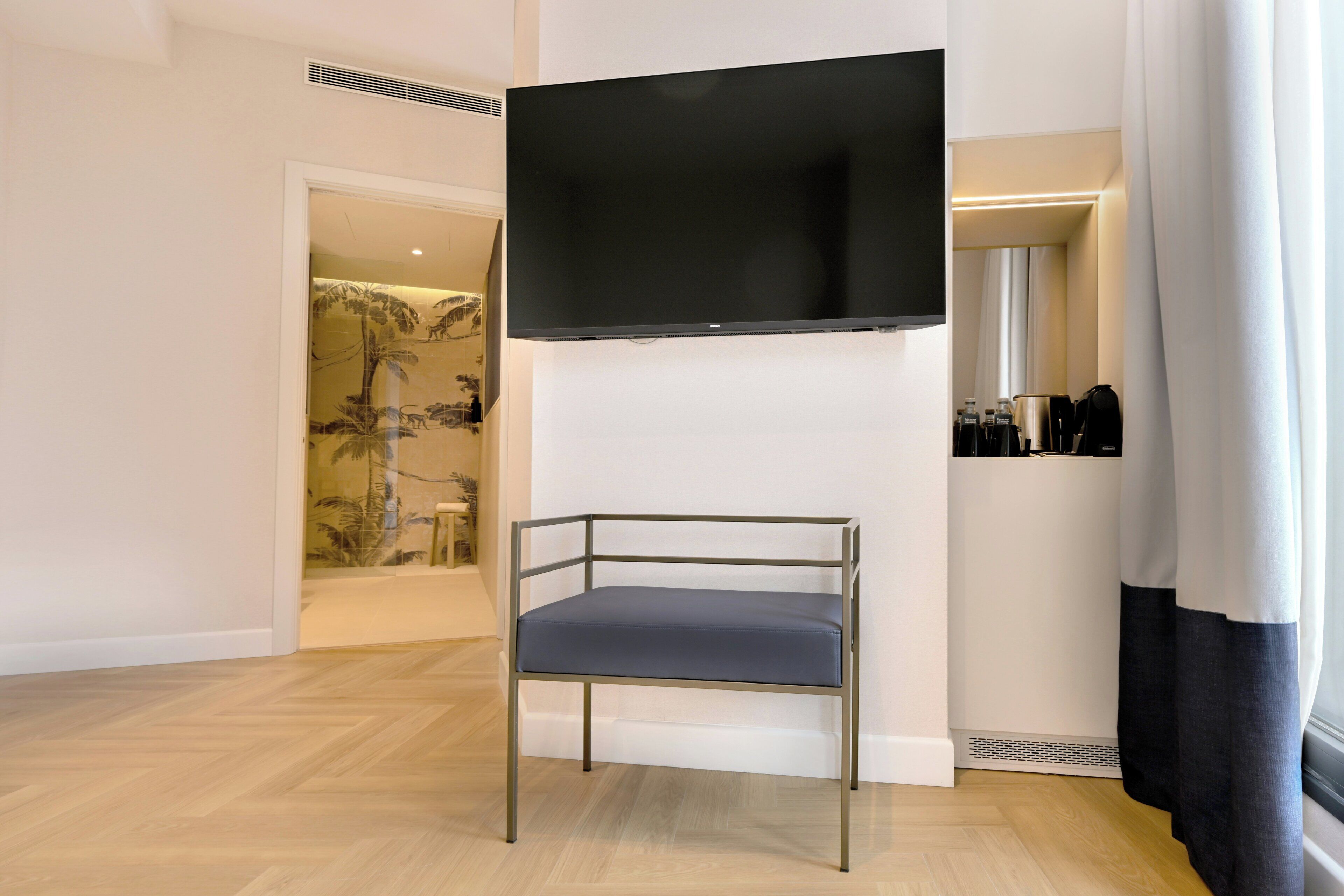 A modern room with a flat-screen TV mounted on the wall, a metal-framed chair with a cushion, and a small shelf with toiletries.
