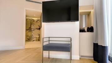A modern room with a flat-screen TV mounted on the wall, a metal-framed chair with a cushion, and a small shelf with toiletries.