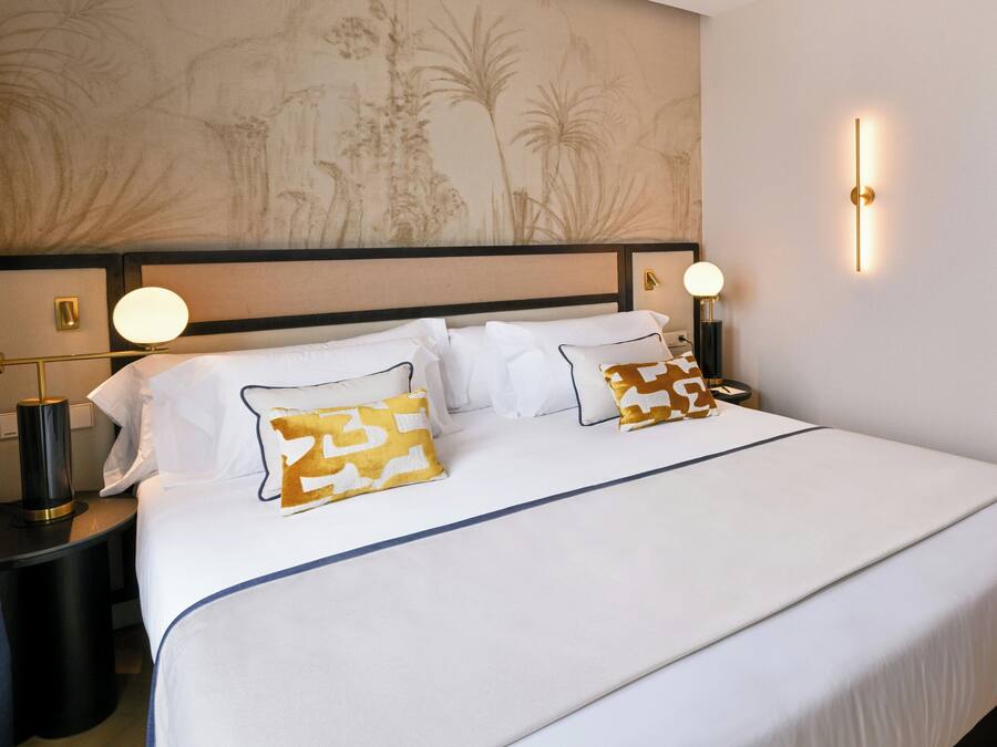 A neatly made bed with white bedding and patterned pillows, a headboard with a floral design, bedside lamps, and a wall-mounted light fixture.
