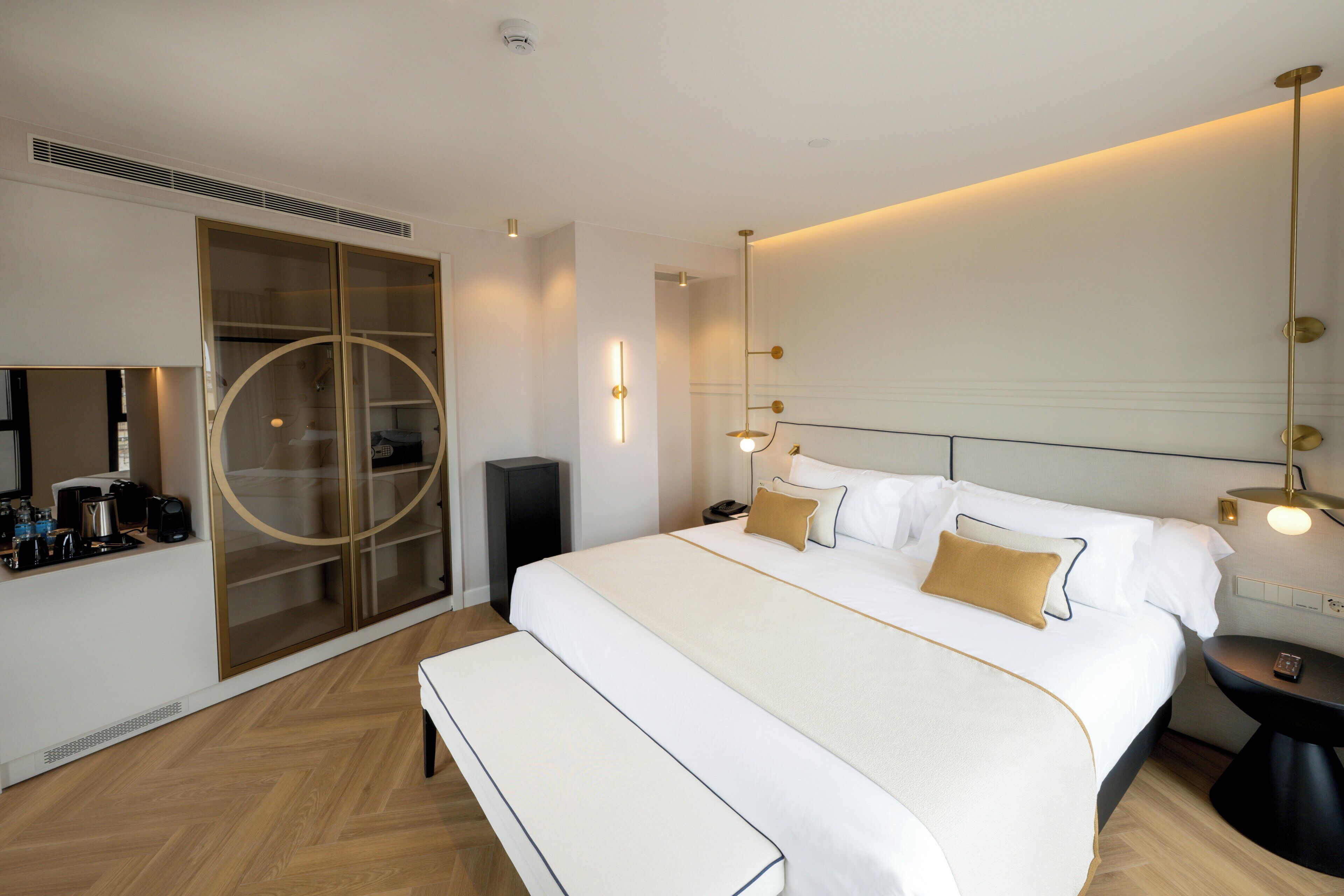 A hotel room with a large bed, a bench, a bedside table, a mirror, a wardrobe, and a television.