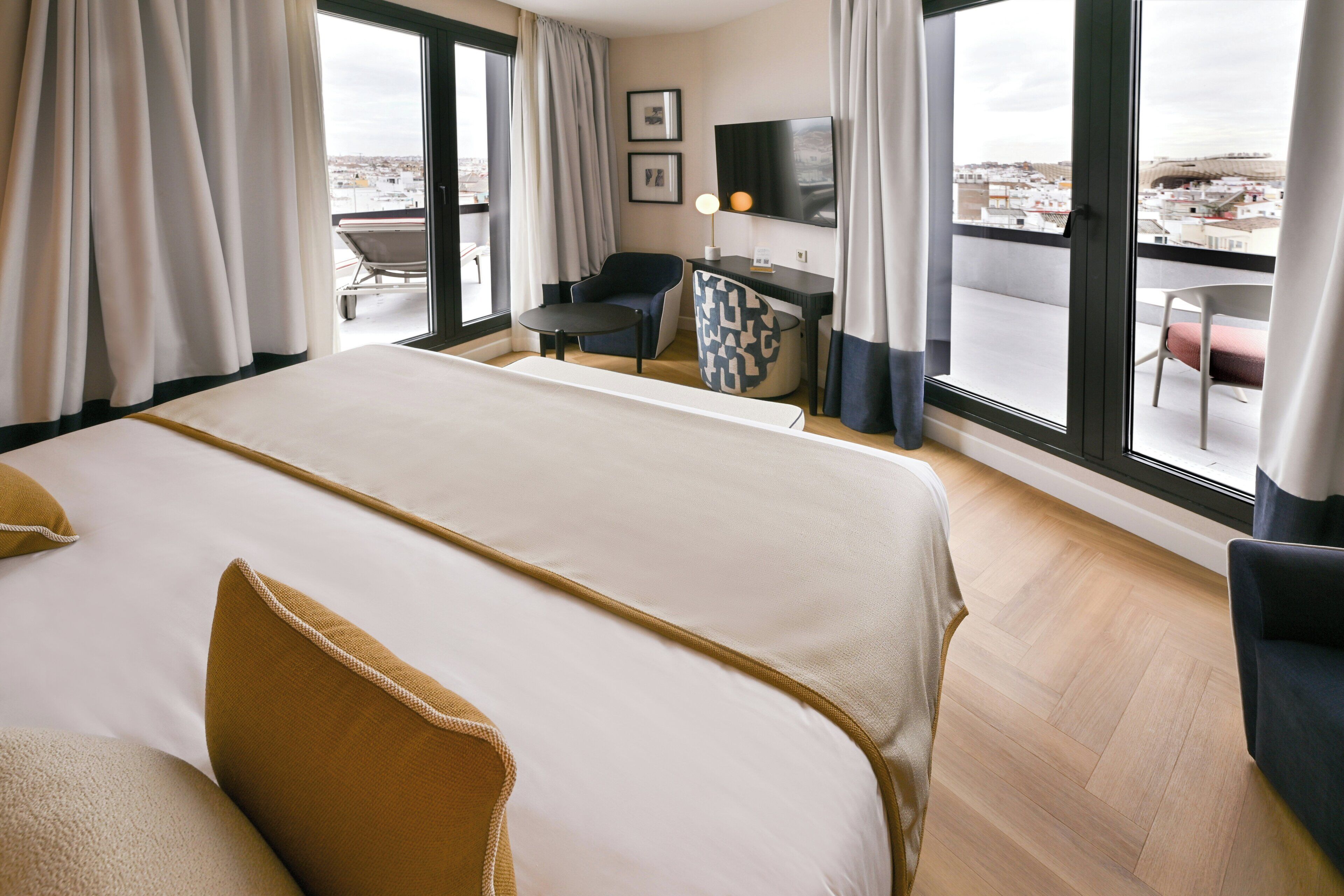 A hotel room with a large bed, a desk with a chair, a TV, and a balcony with a chair.