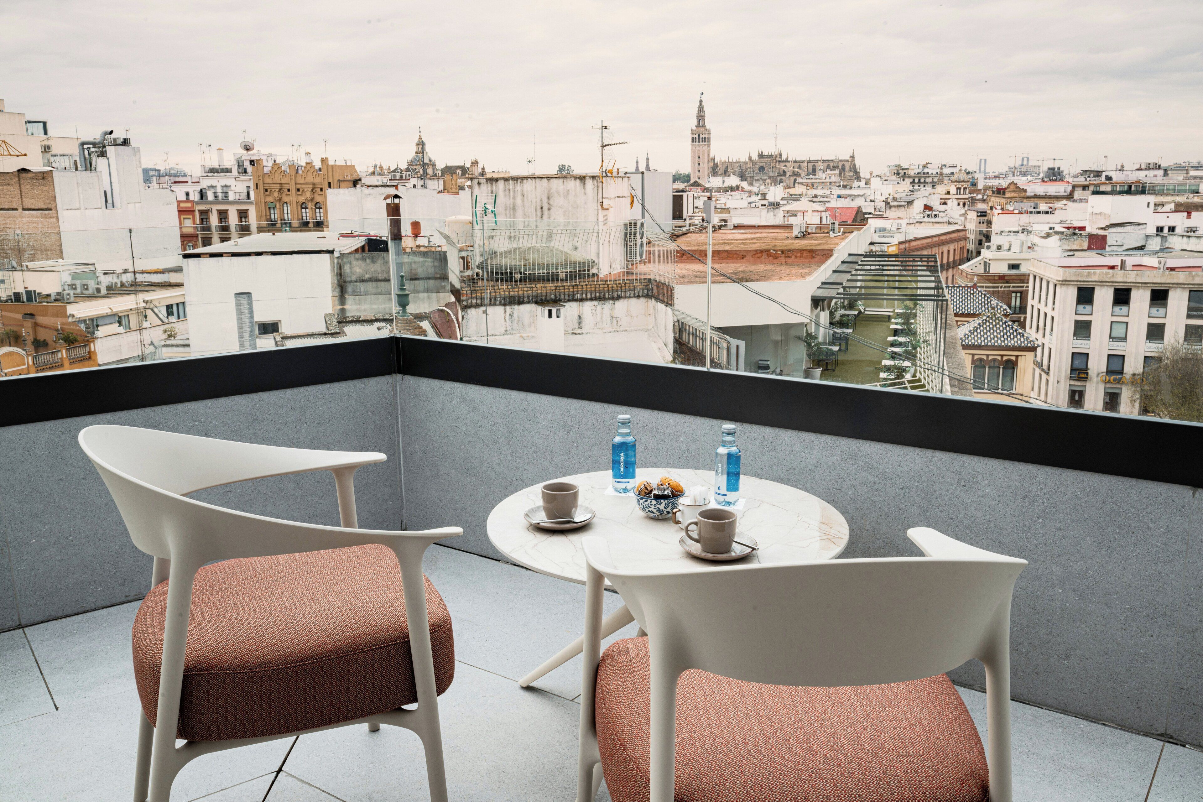 Un balcón con dos sillas blancas y una mesa, vista de un paisaje urbano y un edificio con una torre.