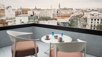 Un balcón con dos sillas blancas y una mesa, vista de un paisaje urbano y un edificio con una torre.
