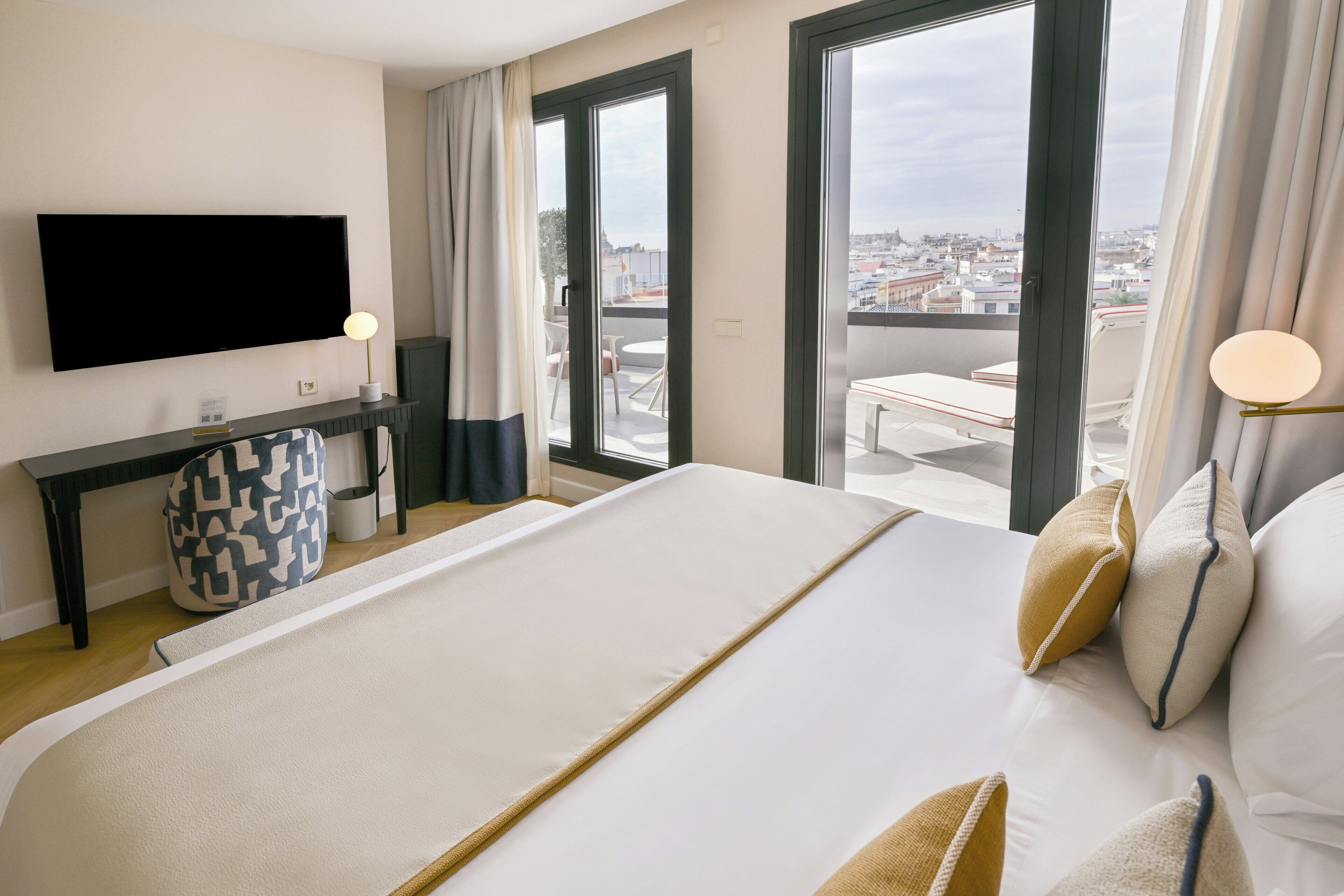 Une chambre d’hôtel dotée d’un grand lit, d’un téléviseur fixé au mur, d’un bureau avec une lampe et offrant une vue sur la ville grâce à de grandes fenêtres.