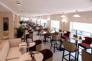 A hotel dining area with multiple tables and chairs, large windows, and ceiling lights.