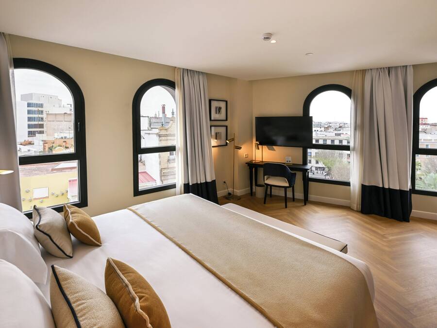 Una habitación de hotel con una cama grande, un escritorio, un televisor y ventanas amplias con cortinas.