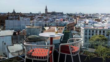Una terraza en la azotea con dos sillones de mimbre blancos y una mesa, con vistas a un paisaje urbano que incluye edificios y una torre.